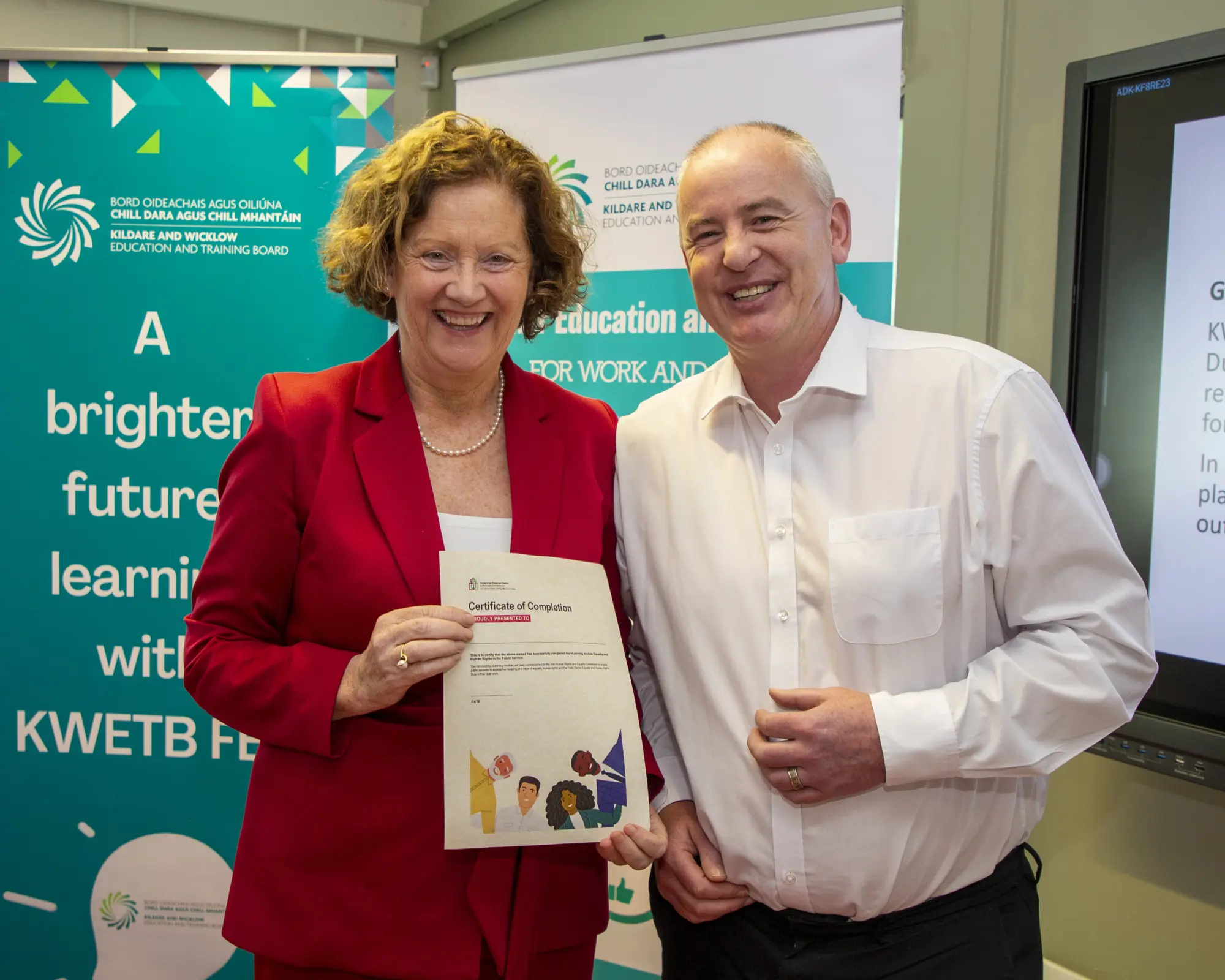 Director of Schools and Chief Executive holding a certificate of completion for IHREC's e-learning module, with KWETB promotional banners in the background.