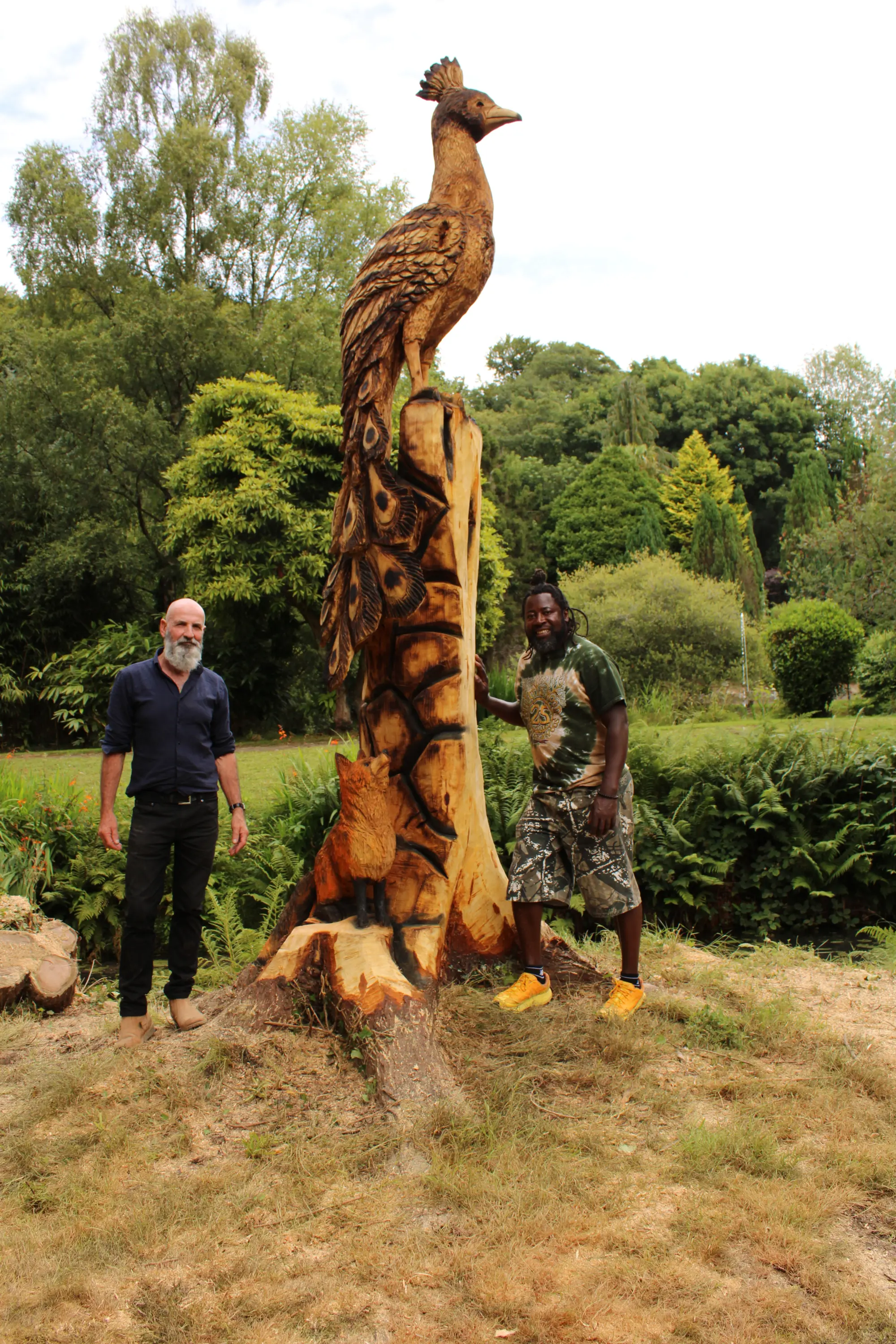 Two men standing beside a tall carved wooden bird sculpture in a garden