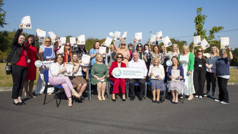 A group of almost 30 persons seated & standing holding up their certificates in celebration, outside in the sunshine