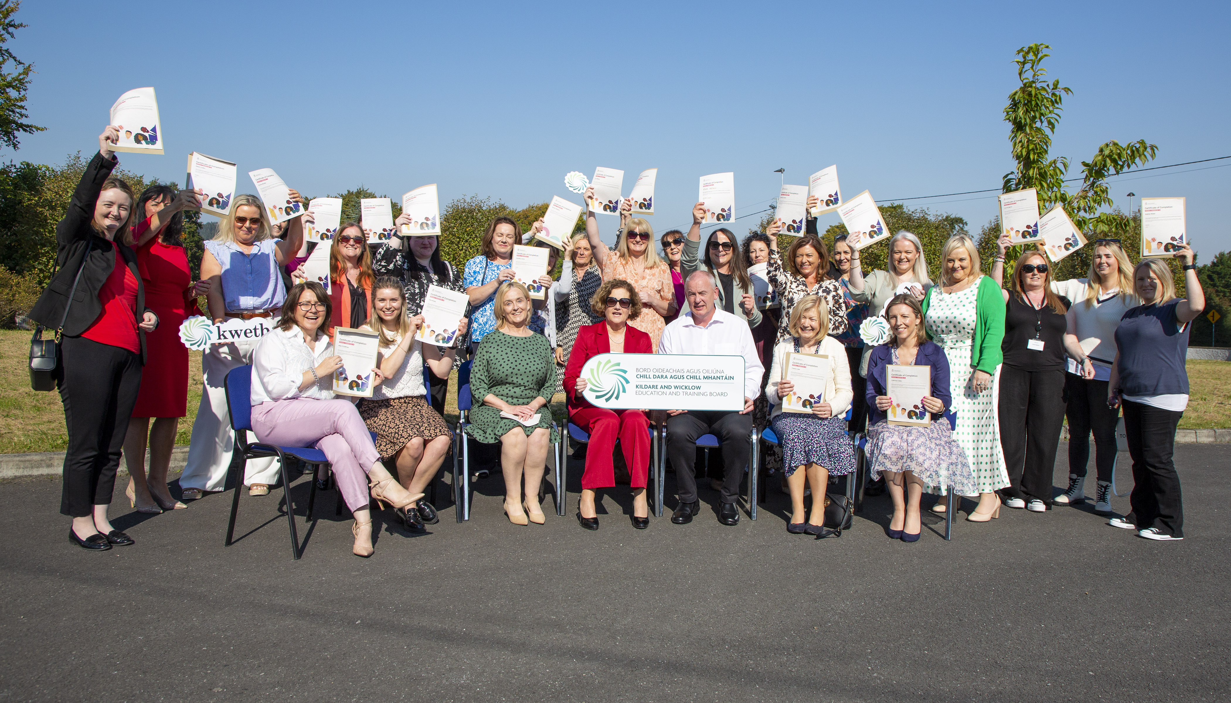 A group of almost 30 persons seated & standing holding up their certificates in celebration, outside in the sunshine